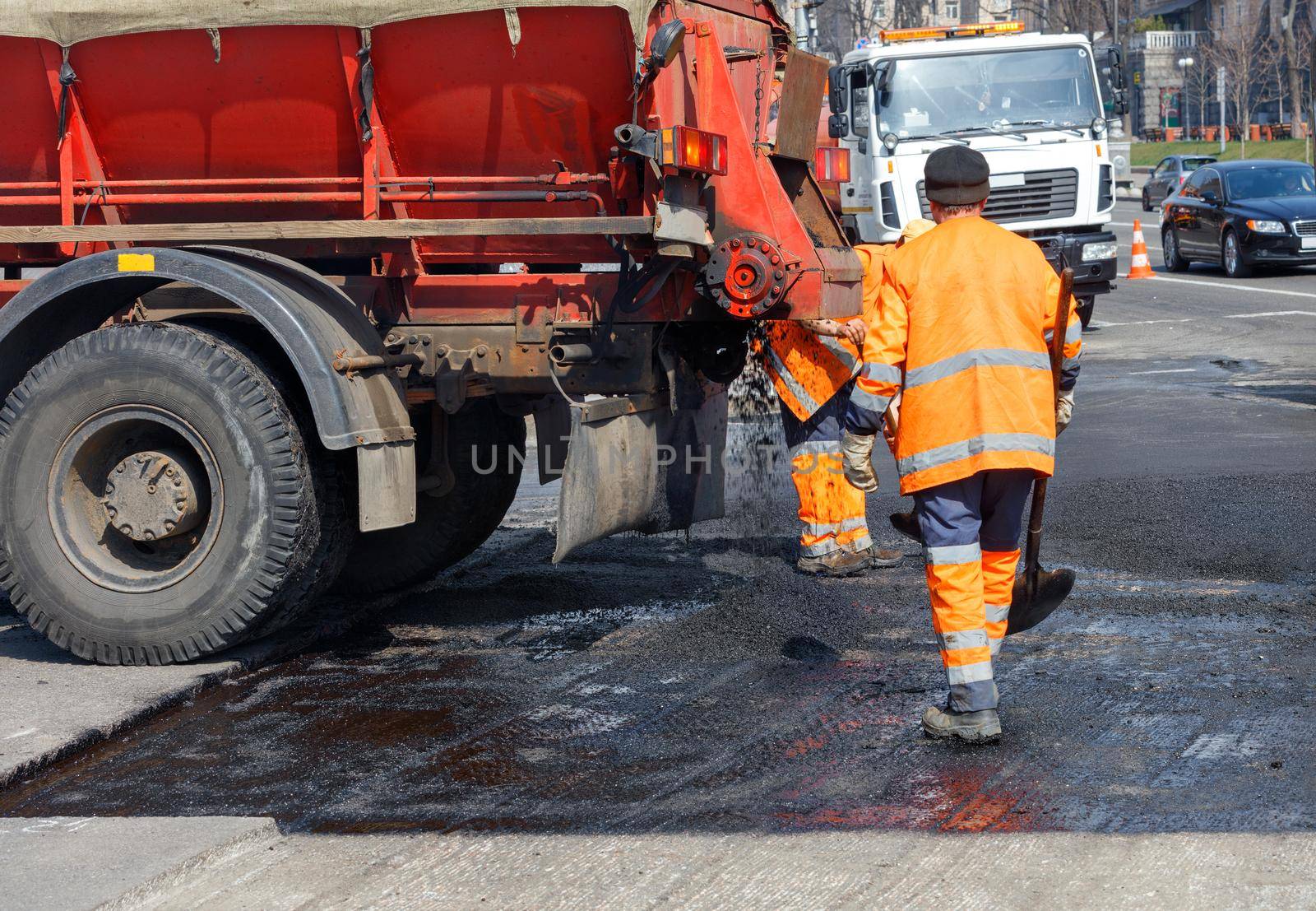 A working group of road workers in orange overalls with a truck for transporting hot asphalt renews part of the road. Road repair concept, place for text, copy space.