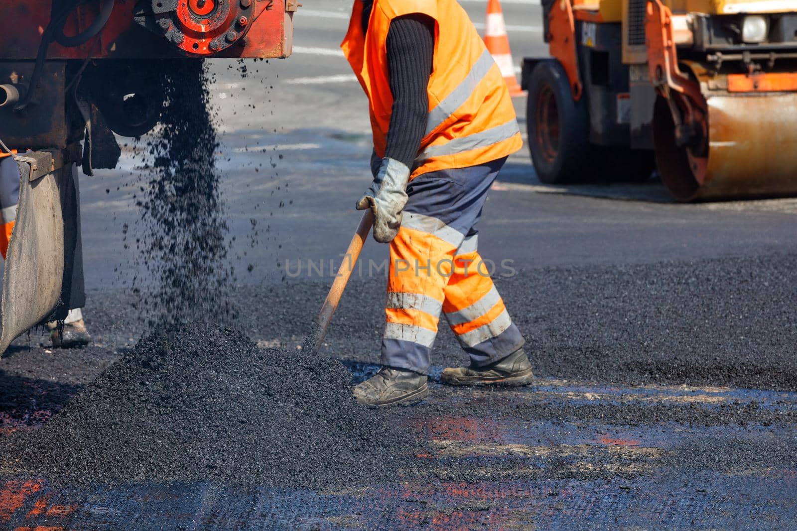 A road worker in orange overalls renovates a section of the road with hot asphalt against the background of a road roller in blur. Road repair concept, place for text, copy space.