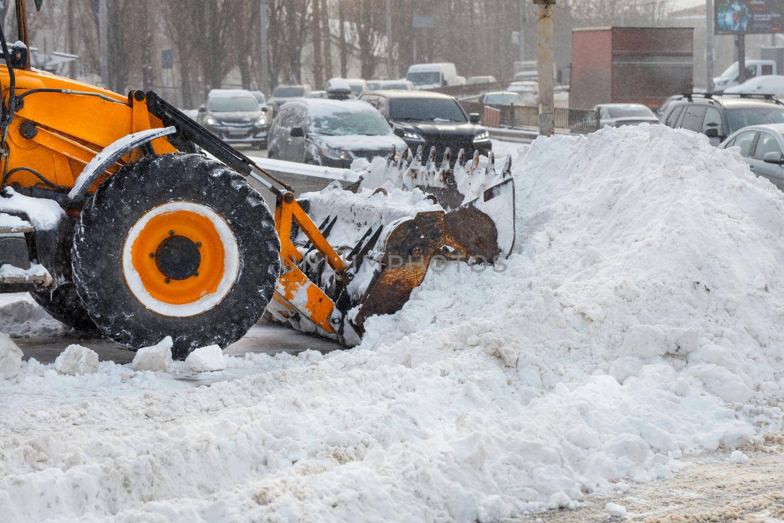 A large ladle of a yellow bulldozer shovels snow from a city road during a snowfall. by Sergii