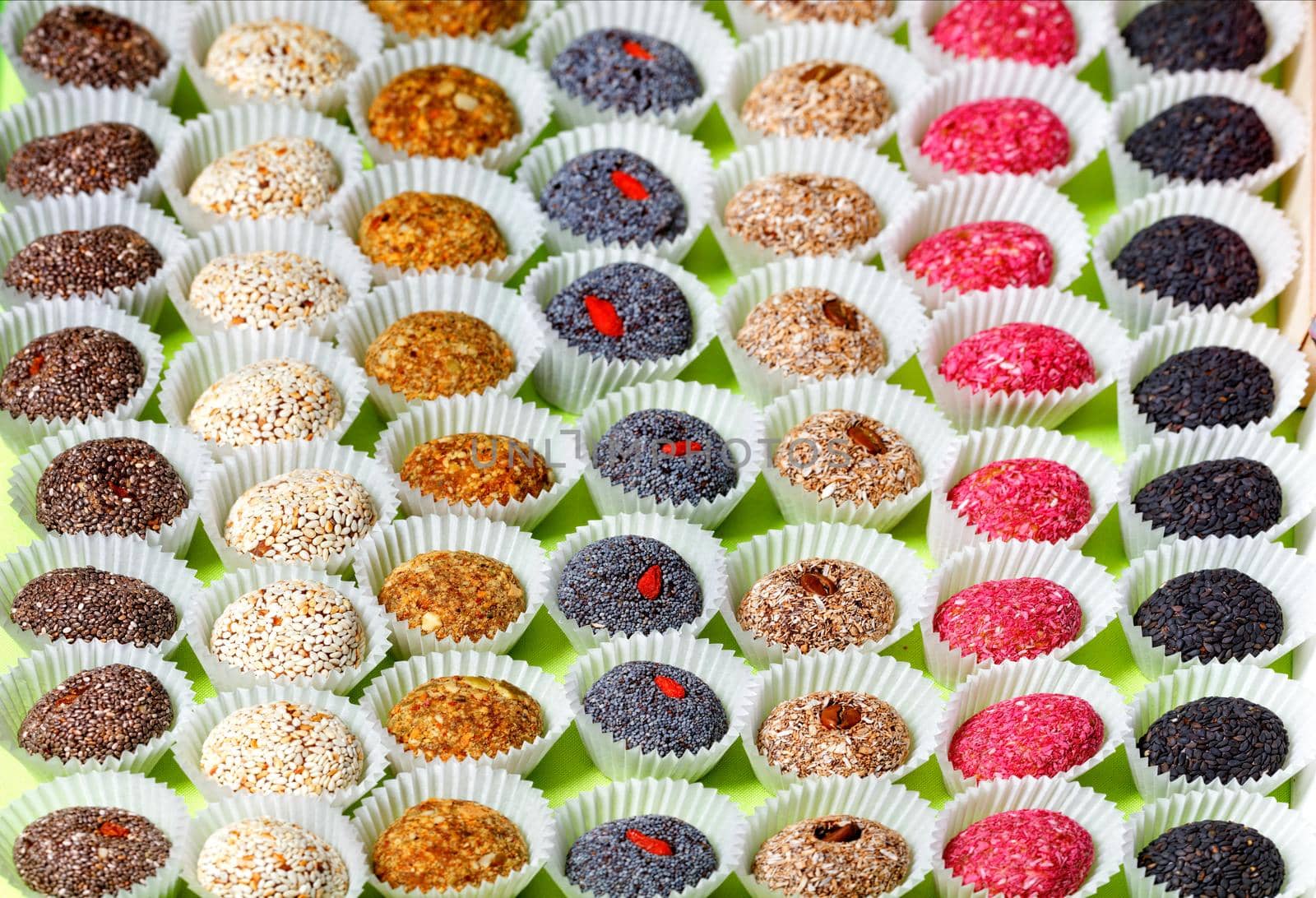 A collection of handmade energy balls with various seed fillings in paper baskets, arranged in even rows. Healthy dessert and appetizer on a bright light green background.