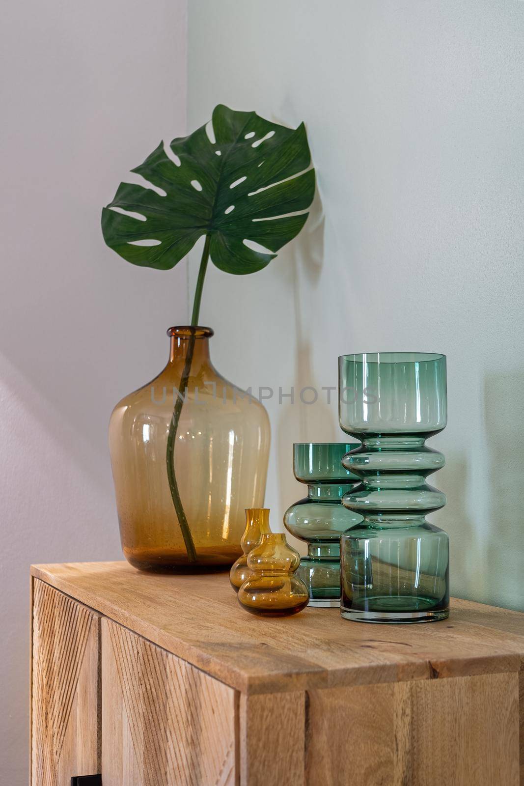 Transparent yellow and green vases for flowers and big bottle with monstera leaf on wooden cupboard. Decoration of interior. by apavlin
