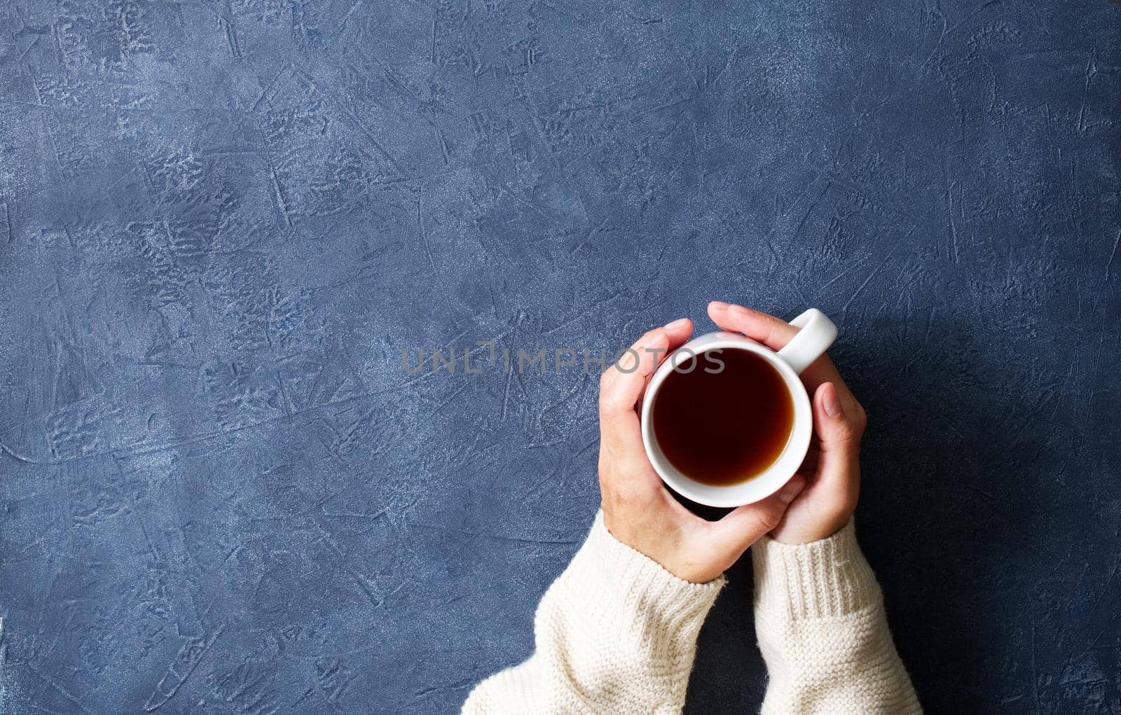 Woman holding cup of hot tea on a dark blue table, hands in warm sweater with mug, winter morning concept, top view