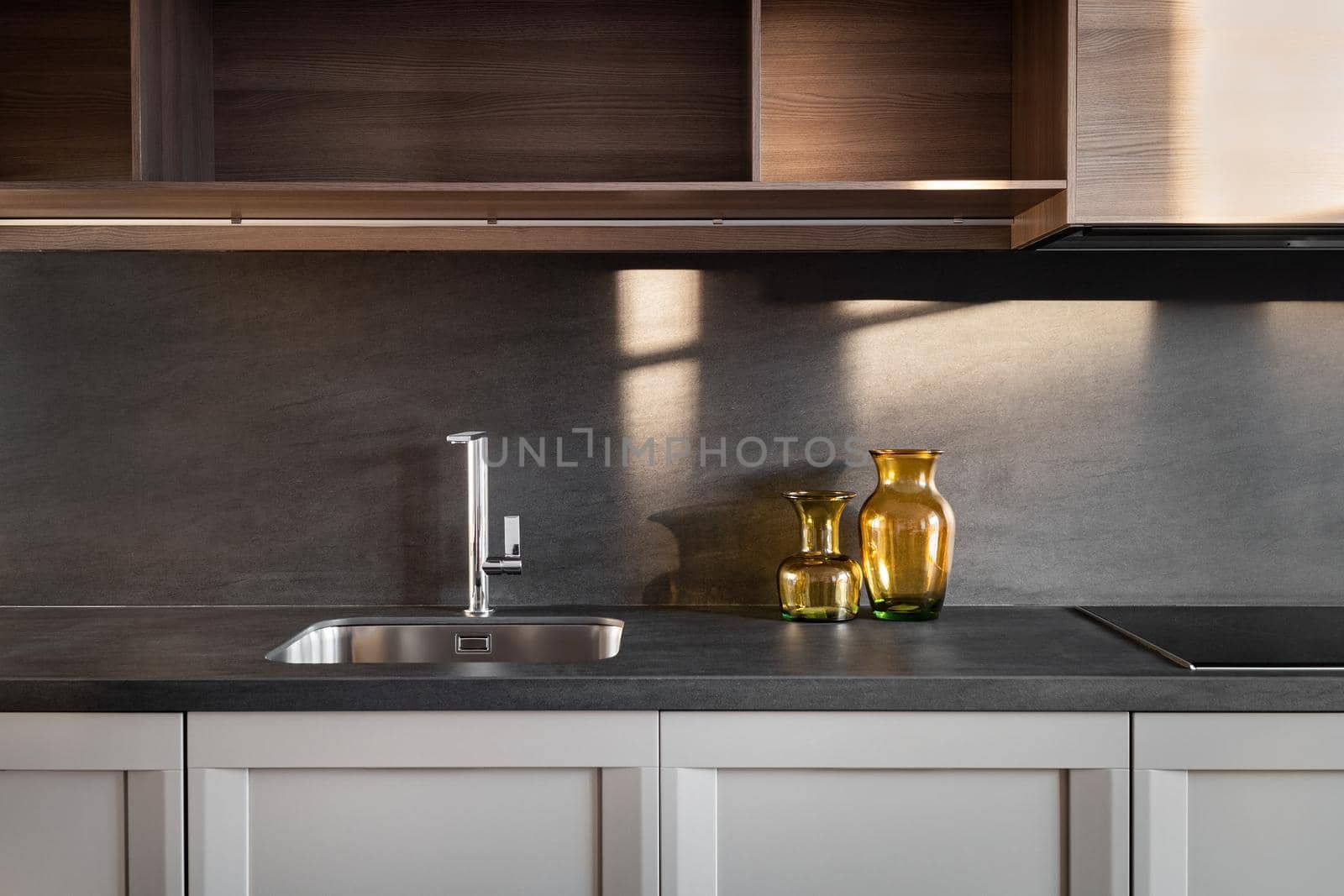 Clean and empty kitchen with wooden cabinets and dark worktop. Steel sink and two transparent yellow, amber vases sunlighted by apavlin