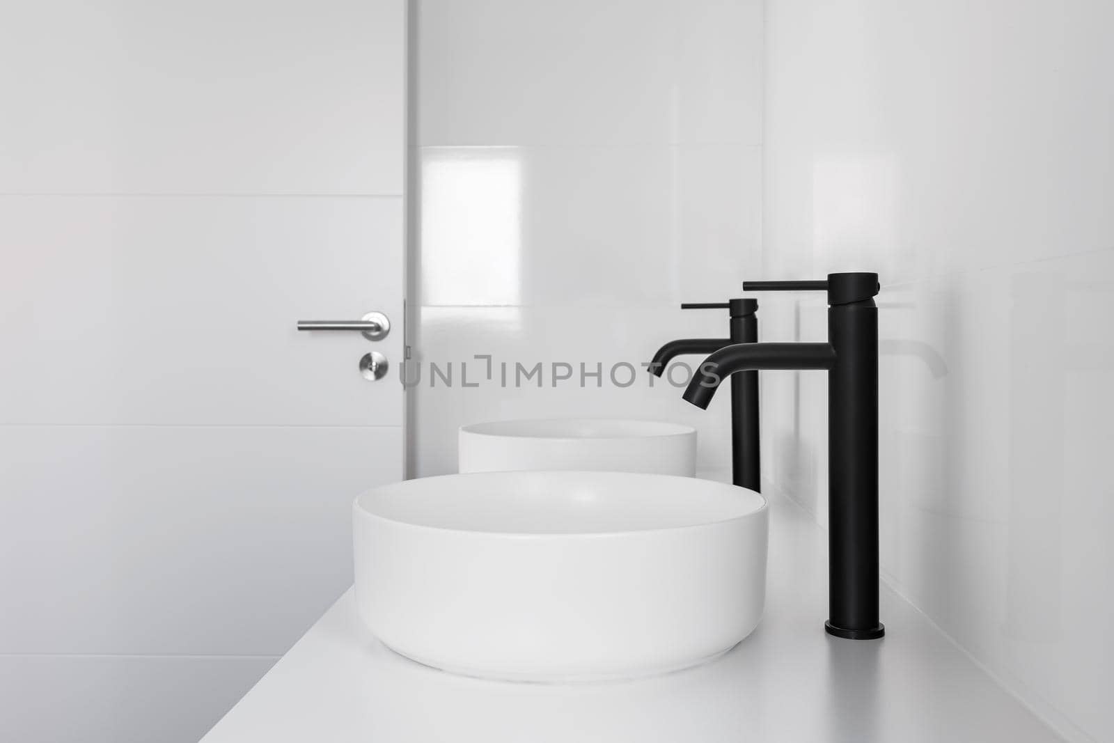 White tiled bathroom with two wash basins and black faucets by apavlin