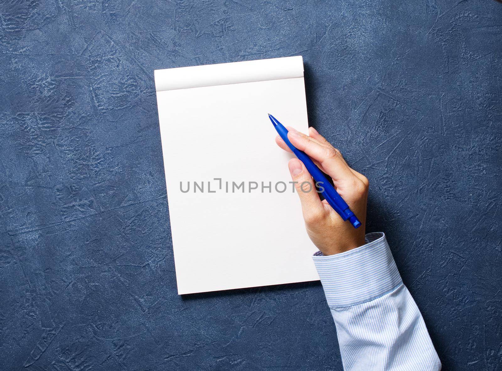 woman writes in notebook on a dark blue table, hand in shirt holding a pencil, sketchbook drawing, top view
