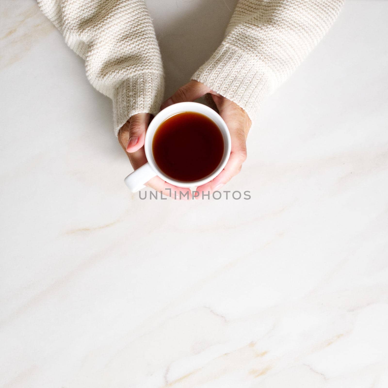 Woman holding cup of hot tea on stone beige marble table, closeup photo of hands in warm sweater with mug, winter morning concept, top view