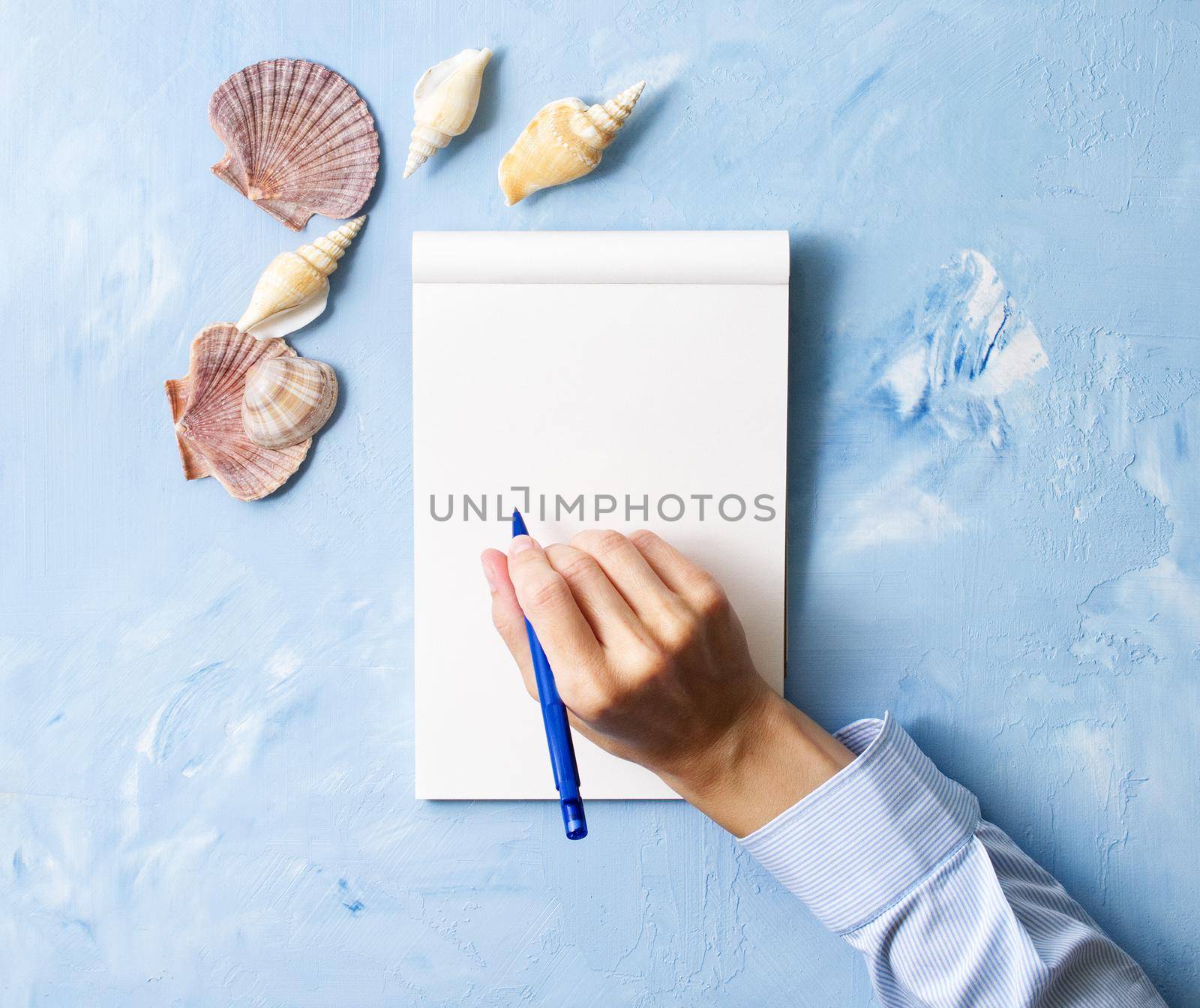 woman writes in notebook on stone blue table, Mock up with frame of seashell, top view, planning holiday by sea by NataBene