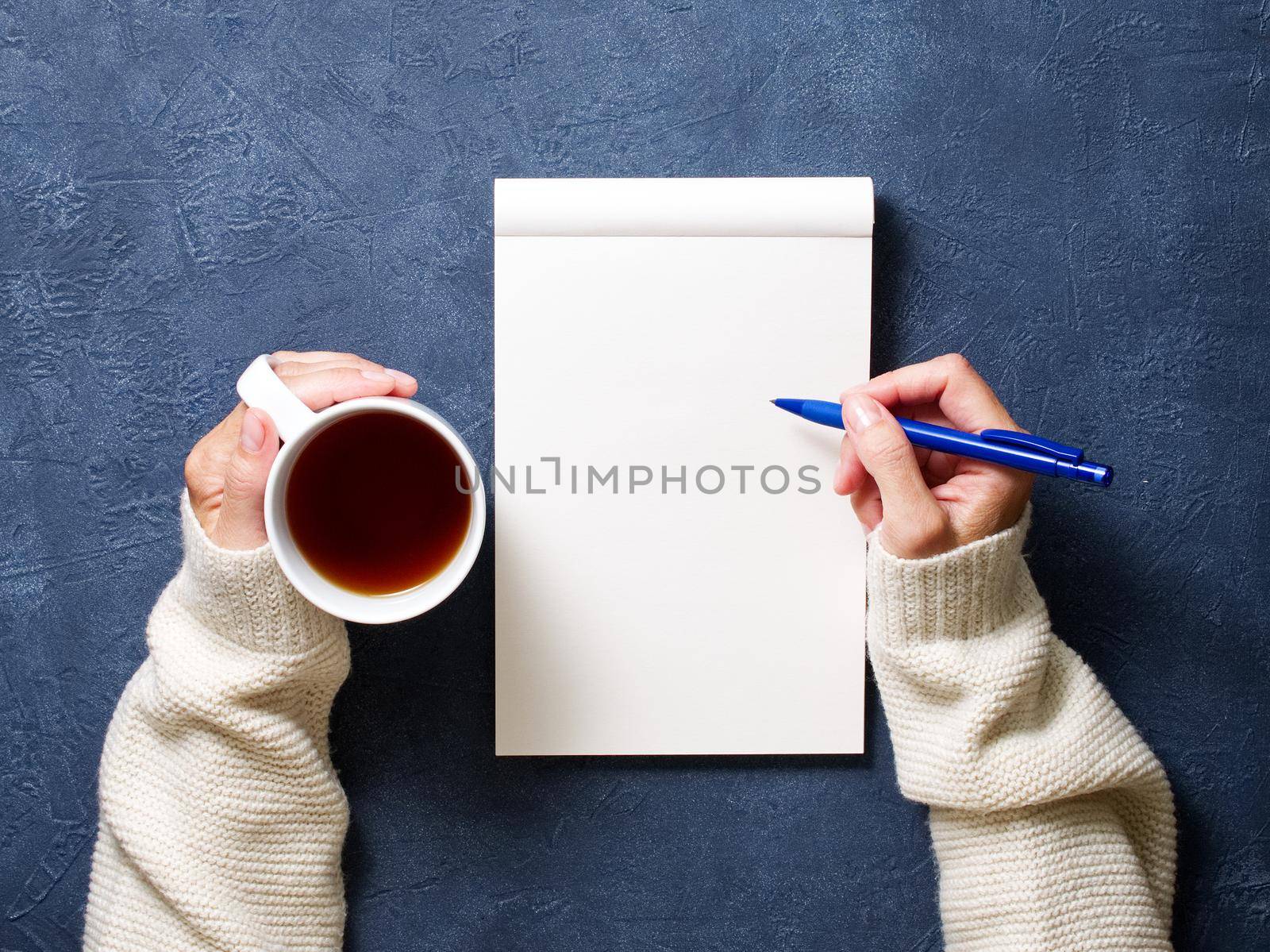 woman writes in notebook on dark blue table, hand in shirt holding a pencil, cup of tea, sketchbook drawing by NataBene