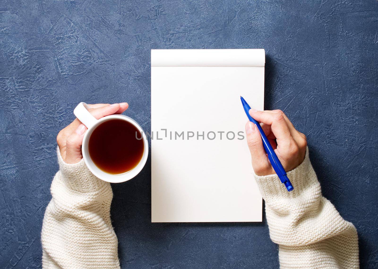 woman writes in notebook on dark blue table, hand in shirt holding a pencil, cup of tea, sketchbook drawing, top view by NataBene