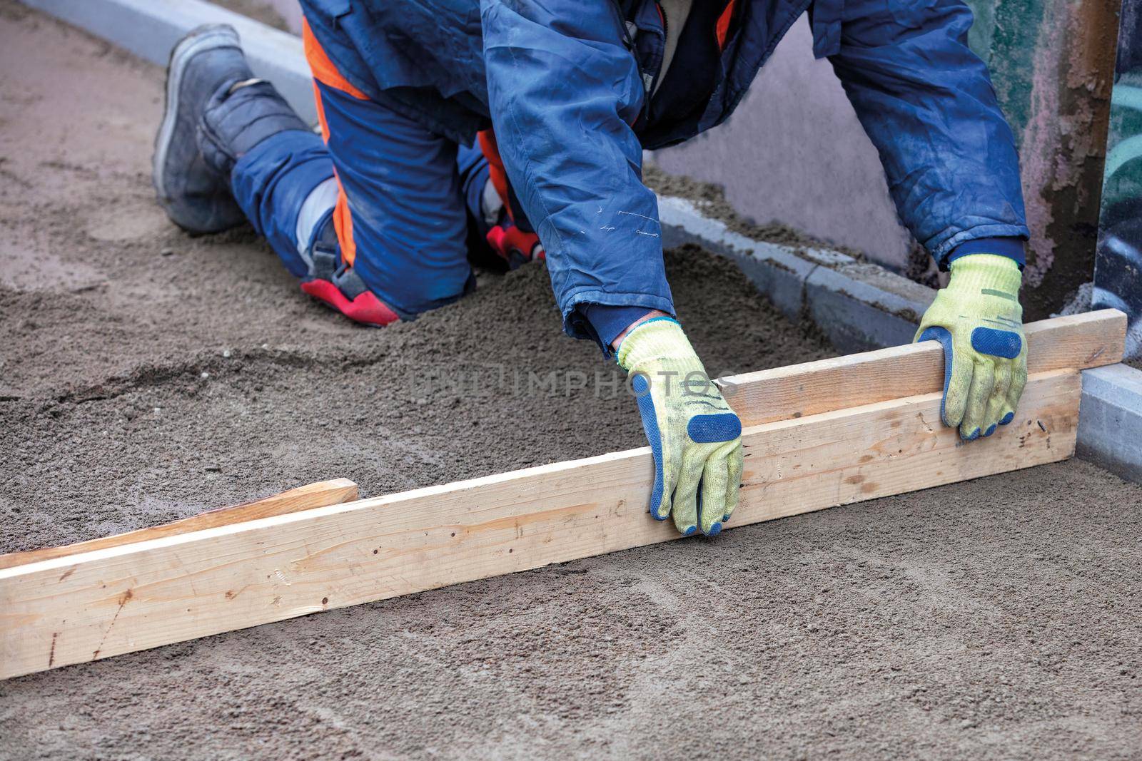 A worker in a blue overalls squats to level the sandy foundation with a wooden level to continue the paving slabs, copy space image, selective focus.