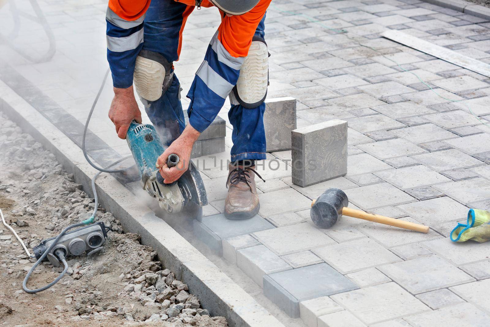 A bricklayer in a blue-orange overalls, bending down, cuts off the protruding part of the paving slabs with a grinder for subsequent even laying. Close-up, selective focus, copy space.