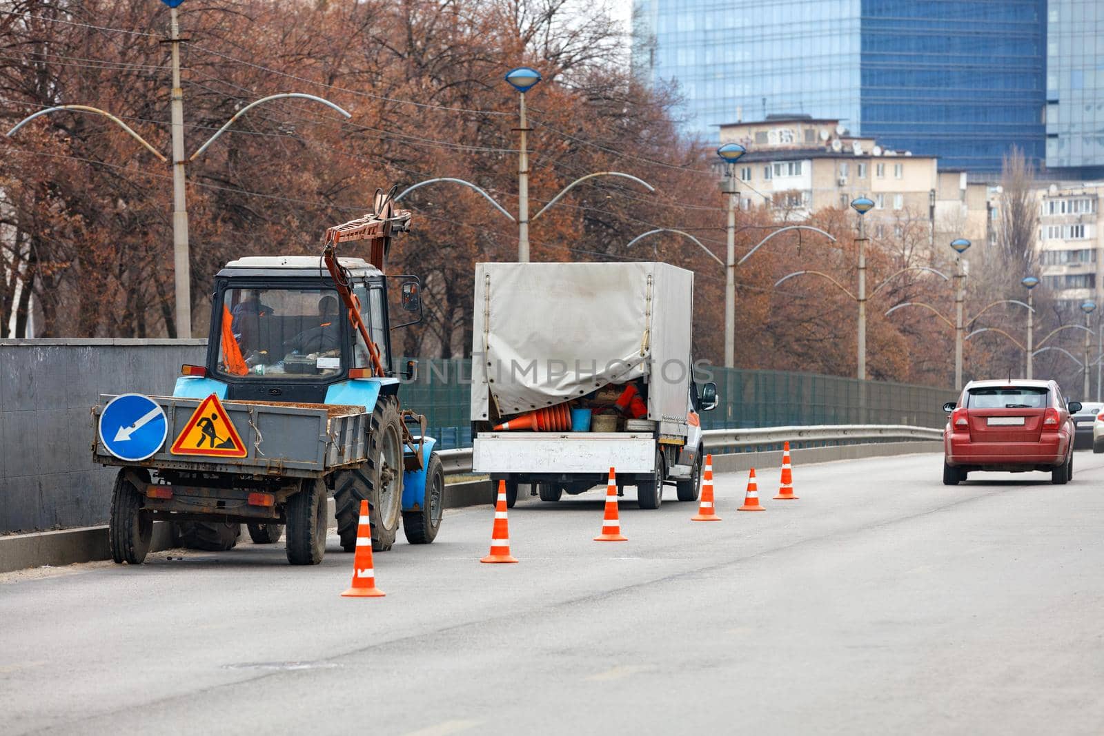 A team of road service workers on a spring day enclose a section of the road with orange cones using an old tractor and truck to clean the side of the road, copy space.