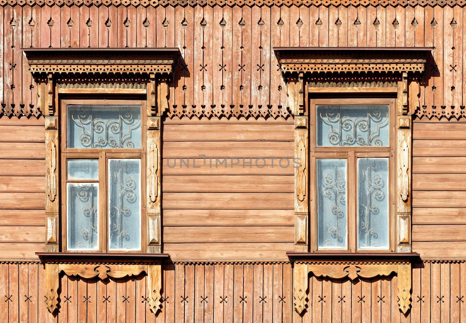 The old wooden textured facade of the house with carved platbands, curly wall elements and metal bars in the windows, illuminated by warm sunlight.