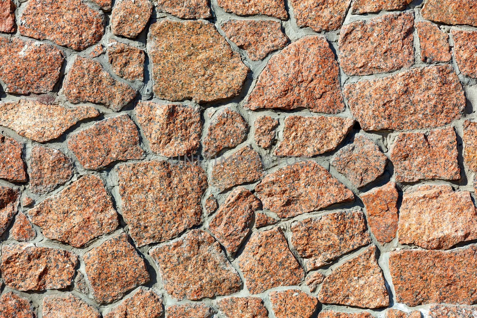 The texture wall of red granite and the background in the form of large blocks embedded in the wall in the light of warm sunlight, close-up, high resolution.