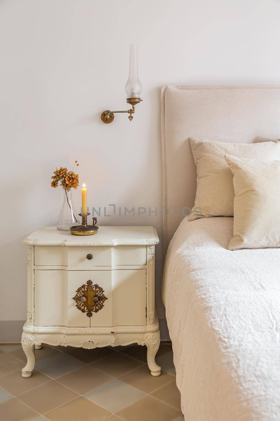 Classic bedroom with wooden bedside table with burning candle and flowers near comfortable bed. Interior of cozy home in retro style