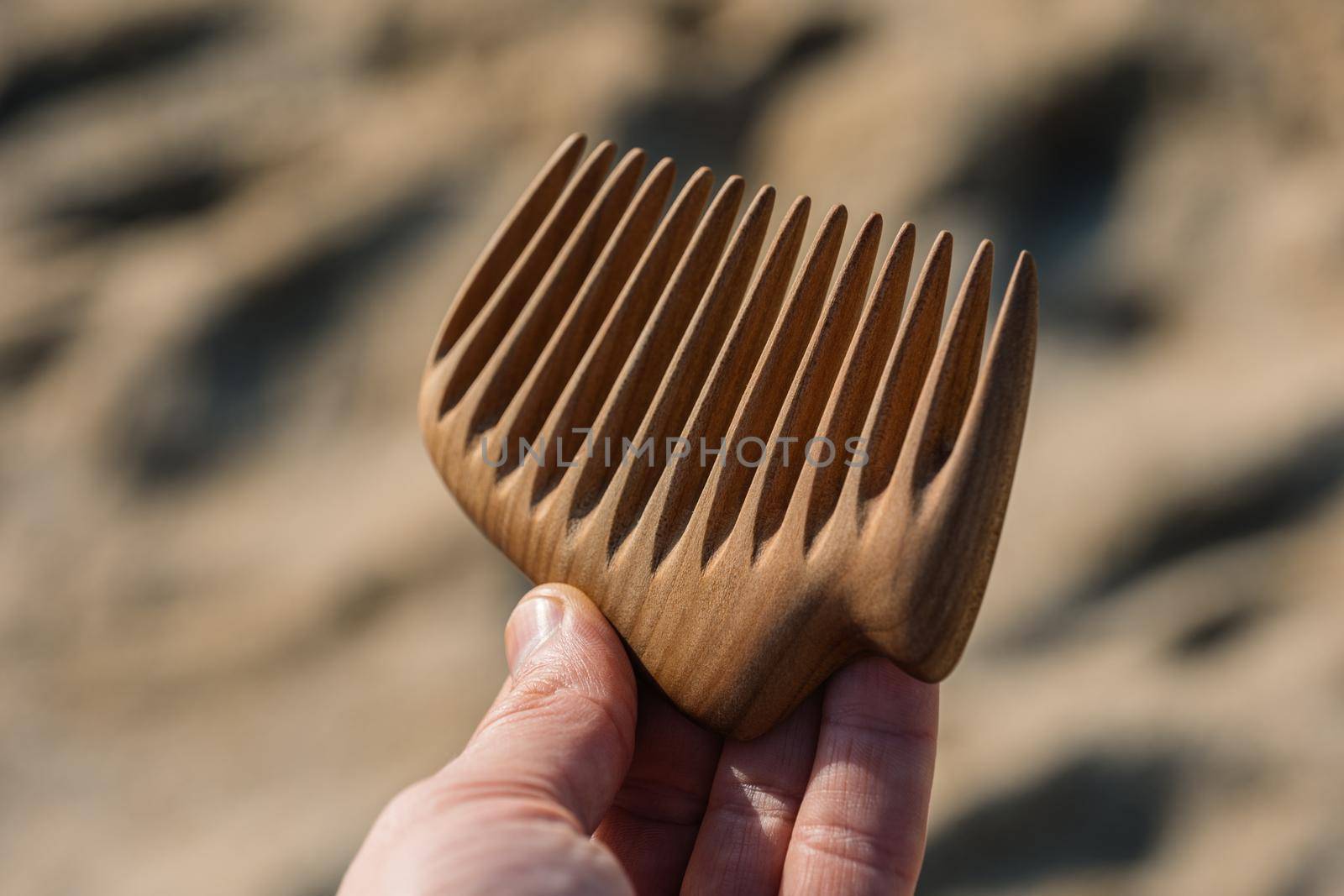 A hand holding natural wooden comb made of pear tree for scalp massage, aroma combing at sand background.