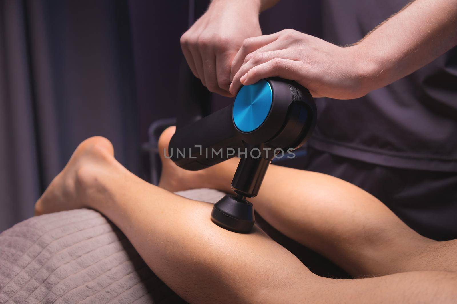 A professional masseur makes a massage of the calf muscle with a massage gun to a man athlete in a massage room with low light by yanik88