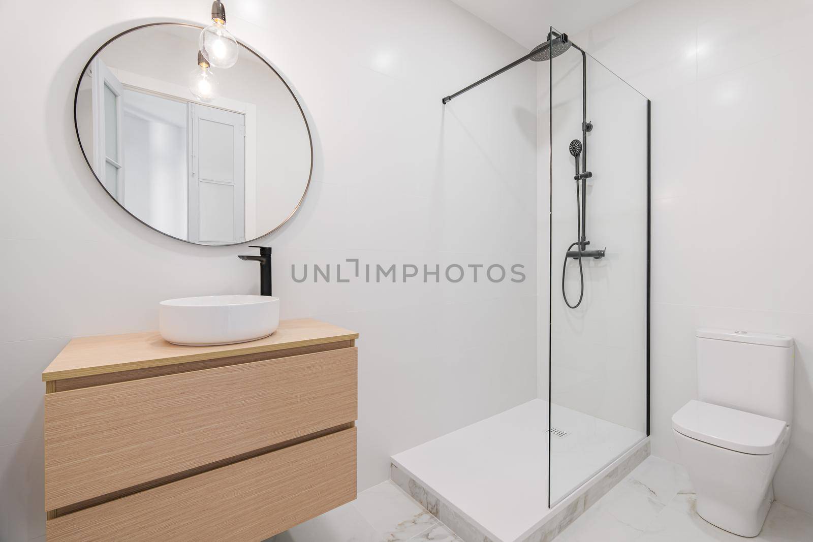 Interior of white bathroom in modern design, with shower zone, toilet, marble floor, wooden furniture and round mirror.