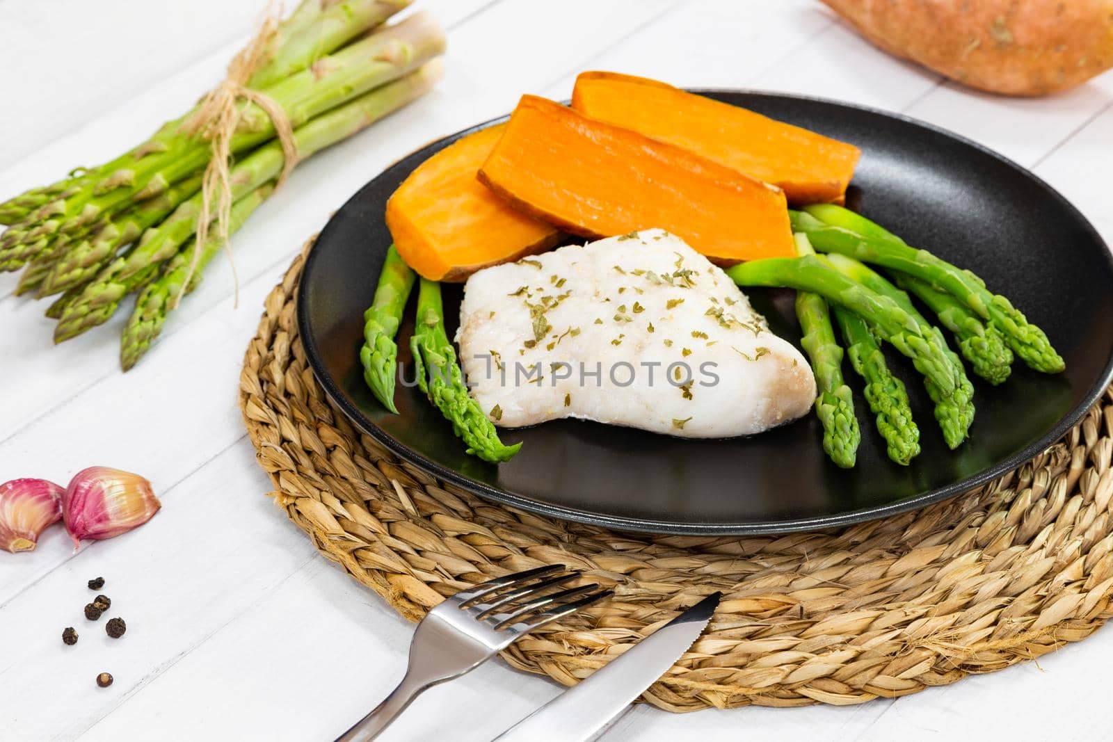 Steamed white fish fillet with asparagus and sweet potato. A portion is served on a black plate in rustic style.