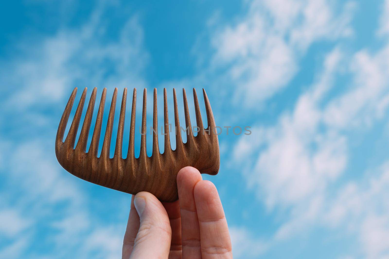 A hand holding natural wooden comb for scalp massage and aroma combing at blue sky background. Hair care concept