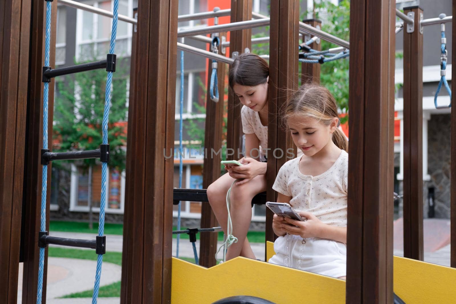 two pretty girls in identical clothes using phone on sports ground or athletic field. kids are surfing in internet. smartphone addiction. advanced, modern children in social networks by oliavesna