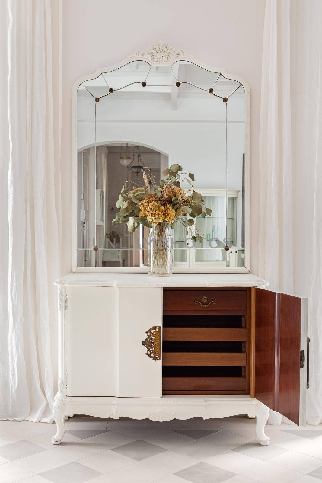 Vintage mirror and flower bouquet on wooden cupboard. Old restored furniture with opened door and drawers in classic bright interior with white wall and curtains by apavlin