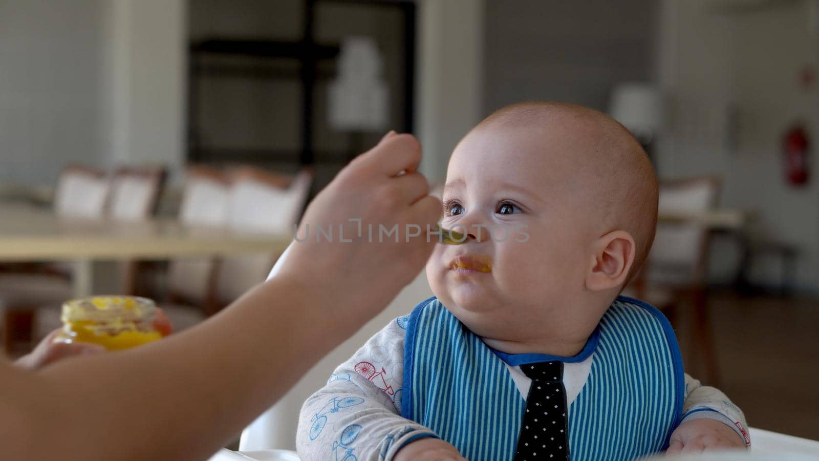mom Mother feed young baby in white feeding up high chair, first supplement vegetable puree Happy smiling kid eat for the frst time, child with dirty face, little infant boy eating porridge nutrition.