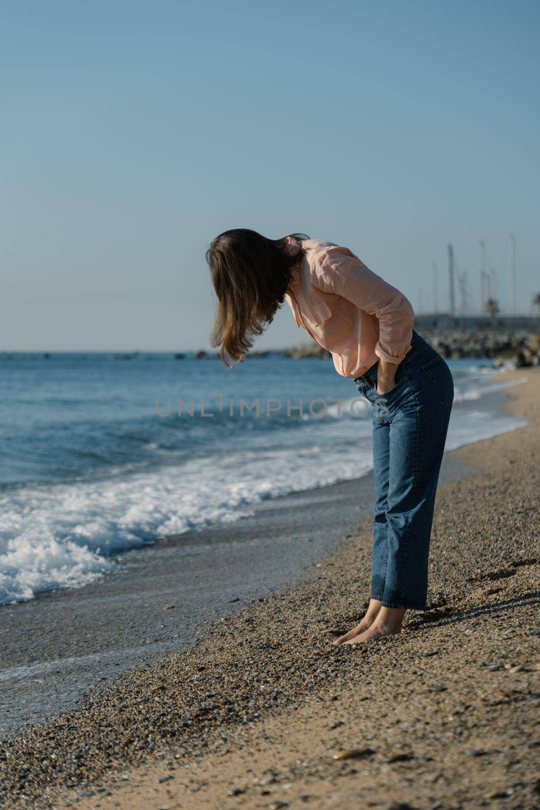 Barefoot woman in pink shirt and blue jeans looks down at her feet on beach by apavlin