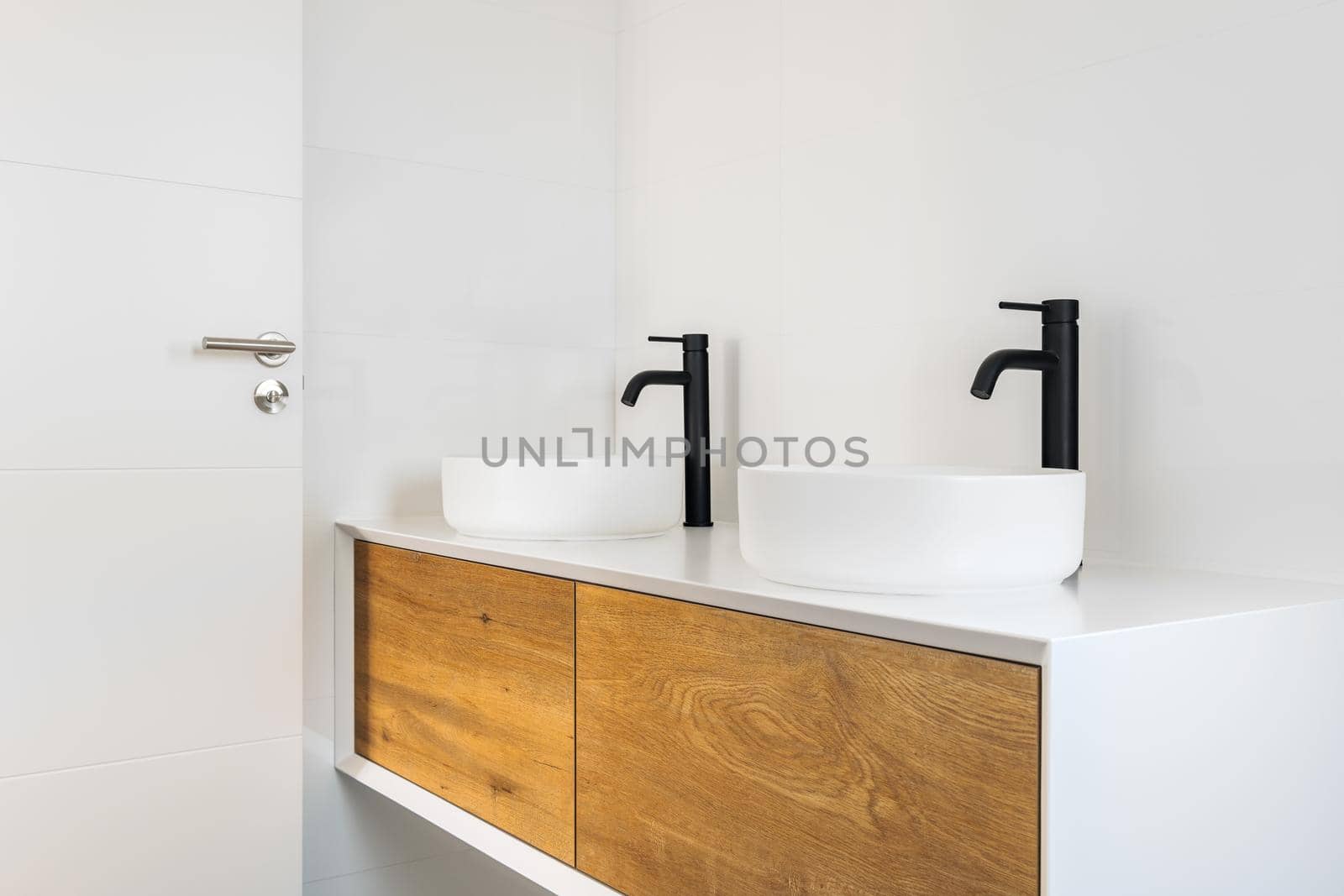 White tiled bathroom with two wash basins, black faucets and wooden cabinets. by apavlin
