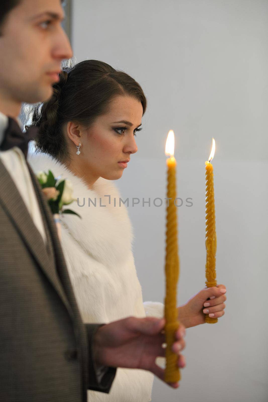 Bride and groom standing at wedding ceremony. Happy stylish wedding couple holding candles with light under golden crowns during holy matrimony in church. by mytrykau