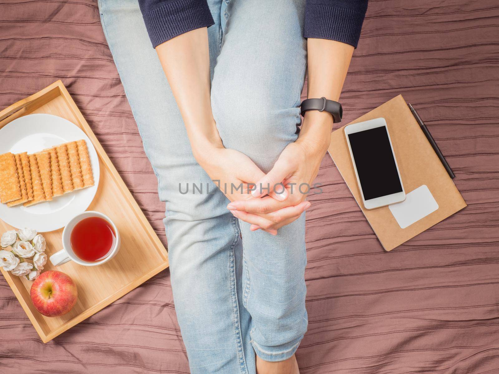 Flatlay of woman in jeans sitting on her bed hugging her knees, with smartphone, notepad, breakfast with tea, top view.