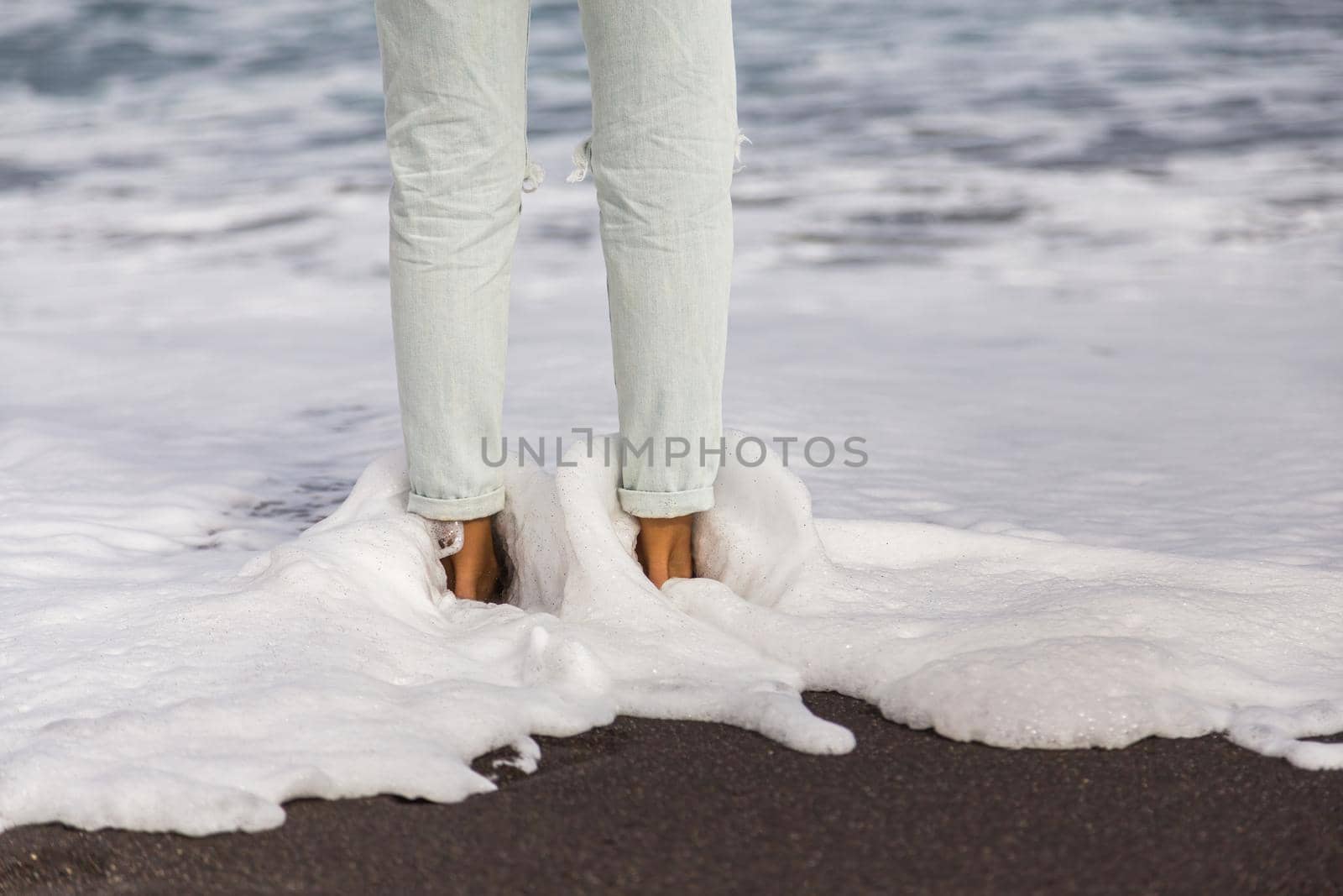 Sea waves with white gentle foam washing over female feet in light blue jeans. Female feet standing on a sand on the beach. Vacation and relax concept.