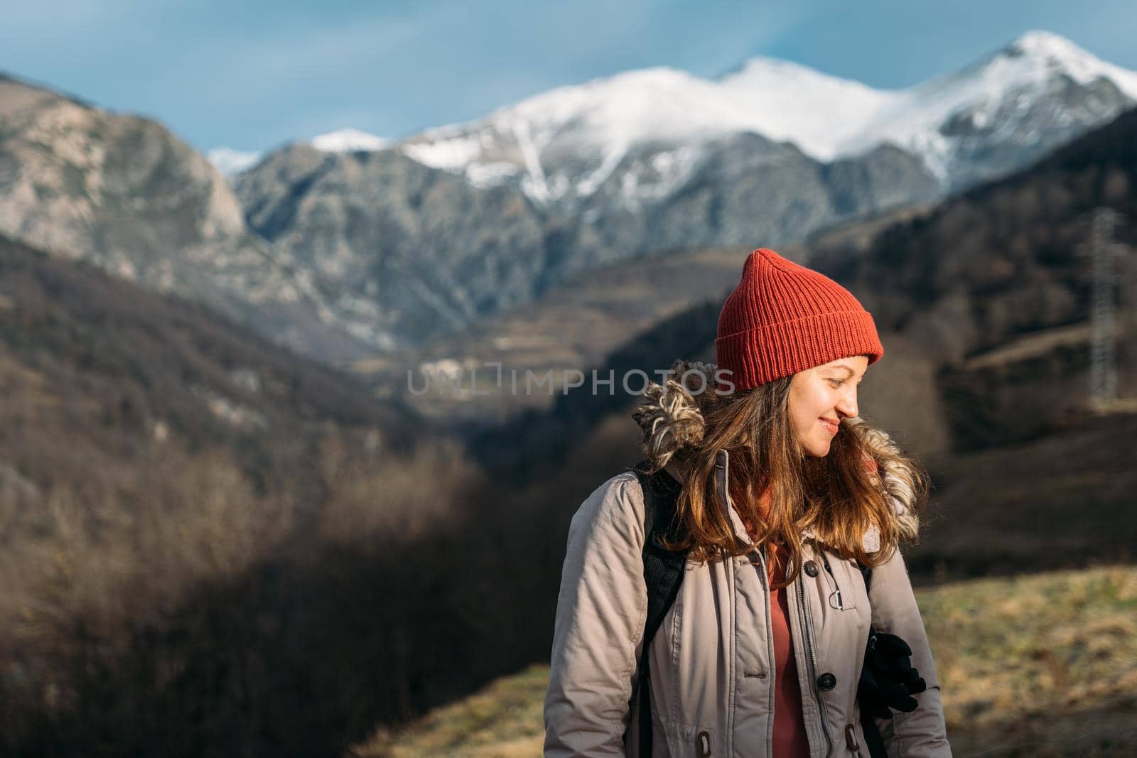 A portrait of a smiling traveler at the snowy mountains background. Happy woman in red cap, warming up and enjoying the sunlight while hiking in the mountains. by apavlin