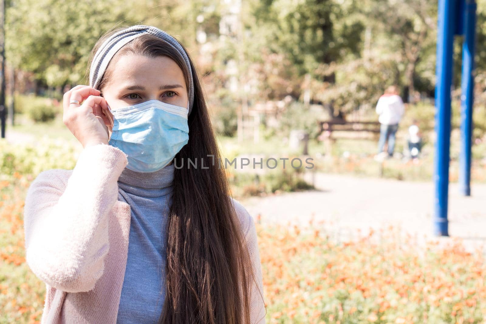 care, infants, spring, coronavirus and quarantine concept - Young cute long haired woman European Caucasian Slavic appearance put on blue medical protective mask in midday sunlight backlight in park