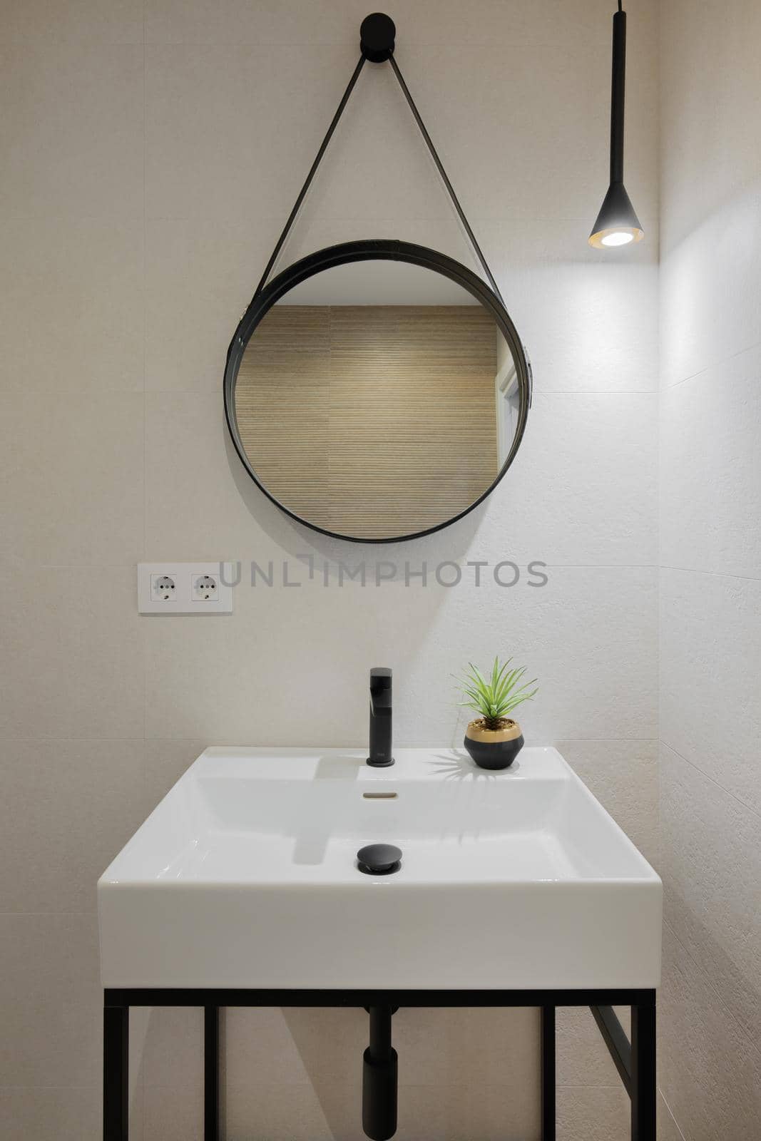 Interior of modern style bathroom in refurbished apartment. White sink with black faucet and round mirror frame, lighted by one lamp