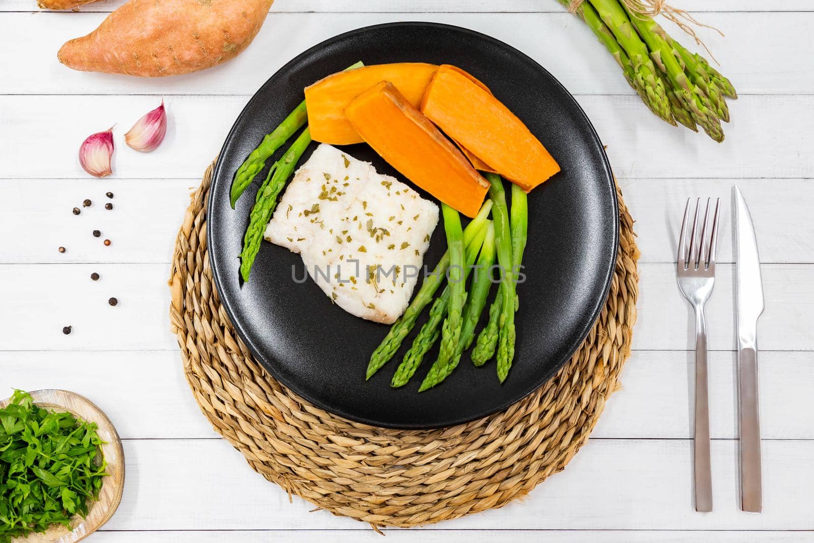 Top view of steamed white fish fillet with asparagus and sweet potato. A portion is served on a black plate in rustic style.