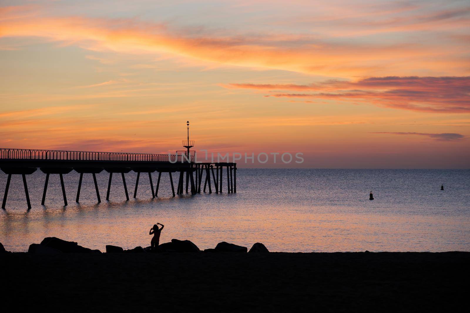 Sunrise in the mediterranean sea with view to the pontoon and silhouette of a woman dancing at the beach in the morning. Pont del Petroli, Badalona, Barcelona, Catalonia, Spain.