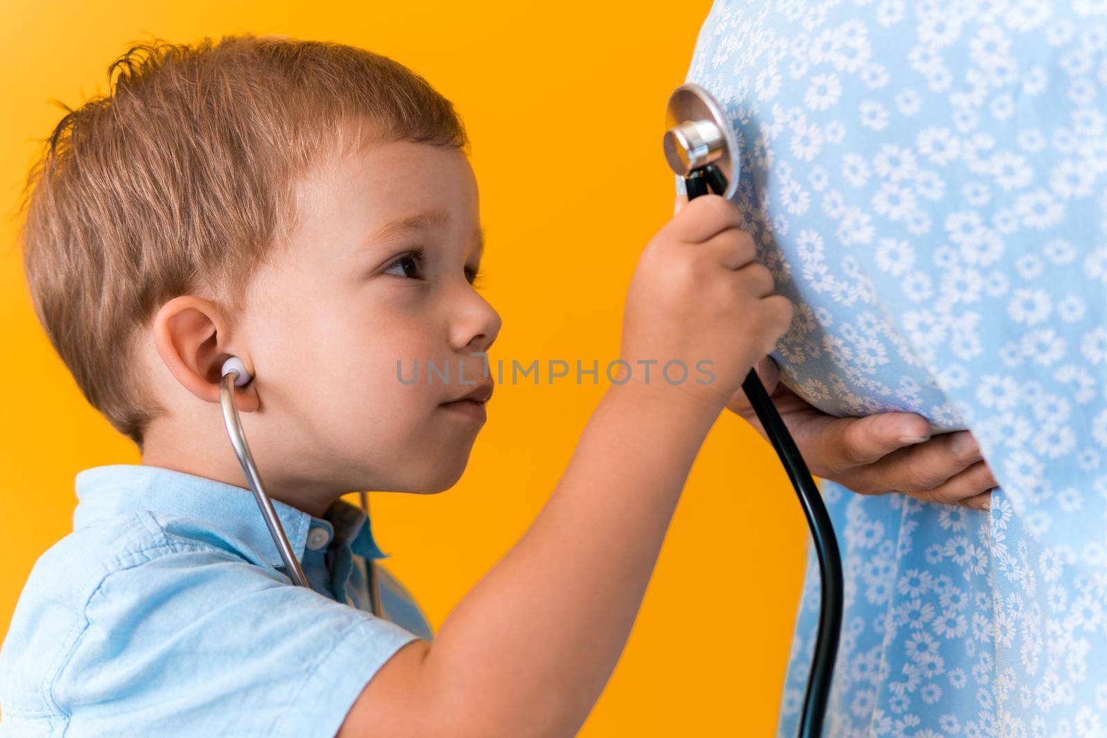 Motherhood, hot summer - croped portrait pregnant mother unrecognizable woman blue dress little boy son sibling treat pregnant mom role play stethoscope unborn brother mom tummy on yellow background.