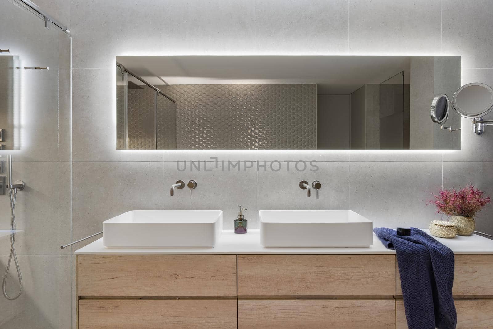 Modern bathroom interior with a wooden shelf, two white sinks and big mirror. Dark blue towel and flowers for decoration