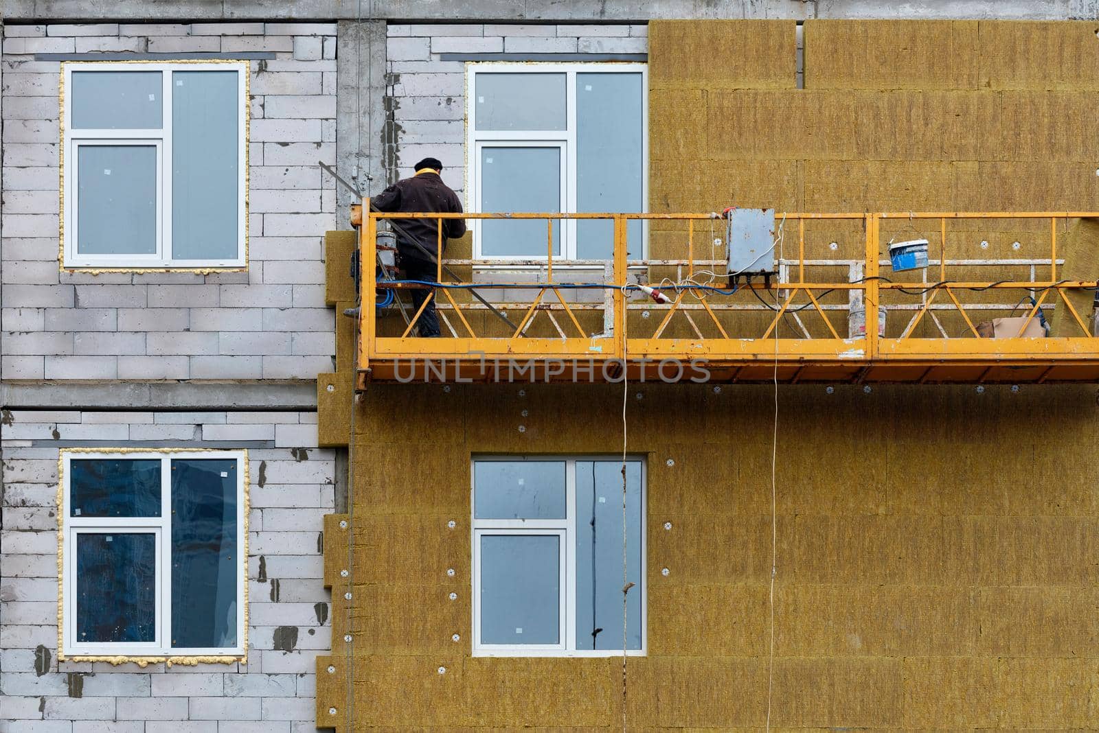 A builder, standing on a suspended platform, insulates the facade of a high-rise building under construction with mineral insulation. by Sergii