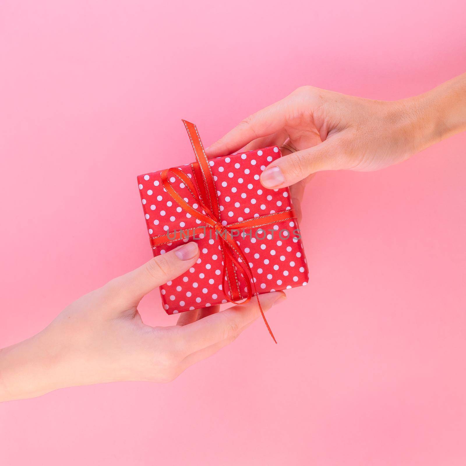 Two women holding a gift in a red gift box with a bow, passing it from one to the other, pink background, top view.