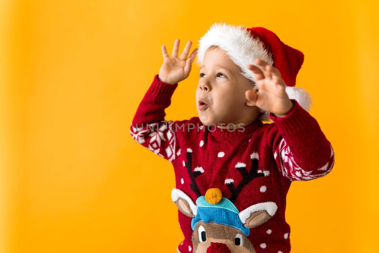 Portrait of happy smiling positive joyful positive preschool little boy in red warm santa hat showing thumb up on orange, yellow background. Winter, holiday, celebration, Christmas, New Year copy space.