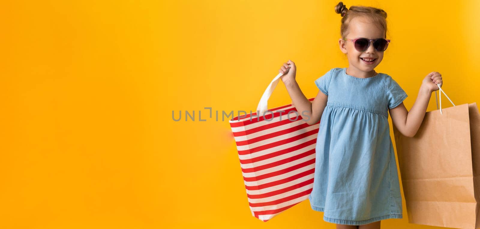 Banner Portrait Beautiful Happy Little Preschool Girl In Sunglasses Smiling Cheerful Holding Cardboard Bags Isolated On Orange Yellow Background. Happiness, Consumerism, Sale People shopping Concept by mytrykau