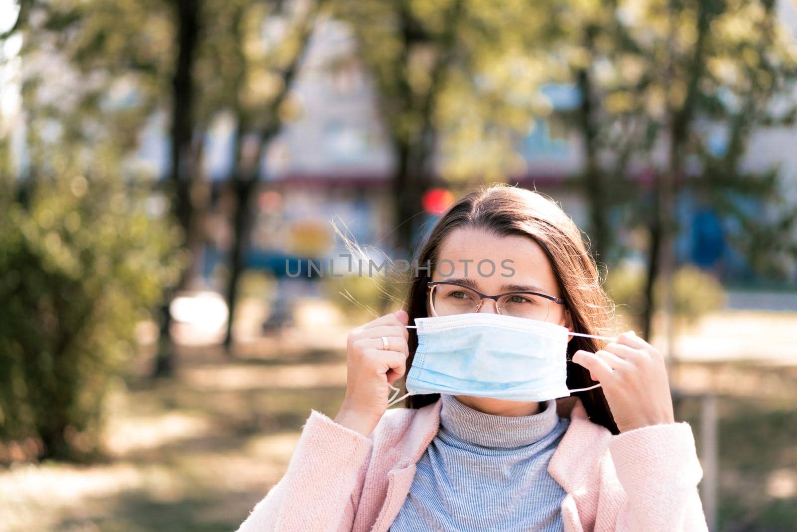 care, infants, spring, coronavirus and quarantine concept - Young cute long haired woman European Caucasian Slavic appearance put on blue medical protective mask in midday sunlight backlight in park