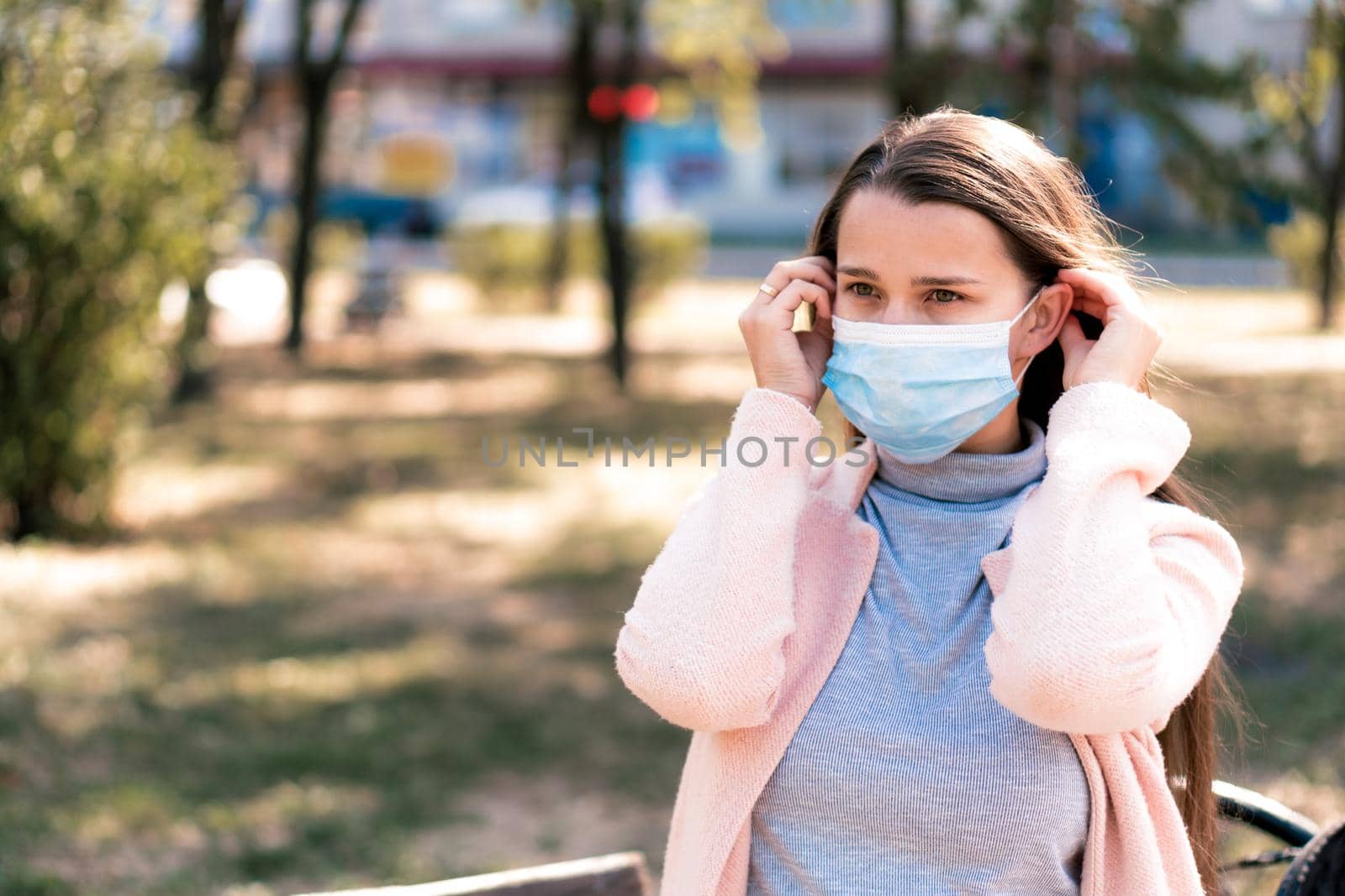 care, infants, spring, coronavirus and quarantine concept - Young cute long haired woman European Caucasian Slavic appearance put on blue medical protective mask in midday sunlight backlight in park