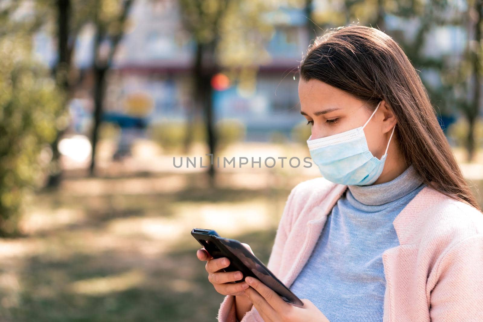 care, infants, spring, cvid-19, quarantine concept - Young cute long haired woman European Caucasian Slavic appearance in medical protective mask uses smartphone in midday sunlight backlight in park.