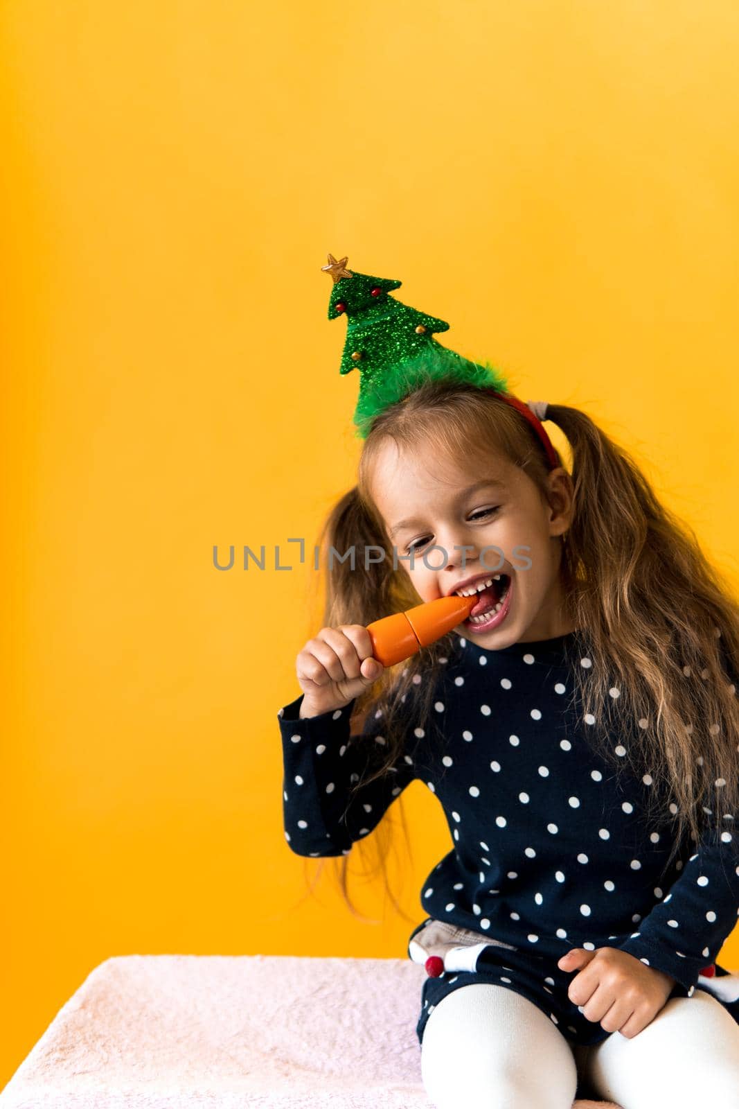 Portrait positive cheerful smiling happy little schoolgirl girl Christmas tree decoration polka dot dress biting eat orange carrots on orange background. New year, holiday, celebration, winter concept.