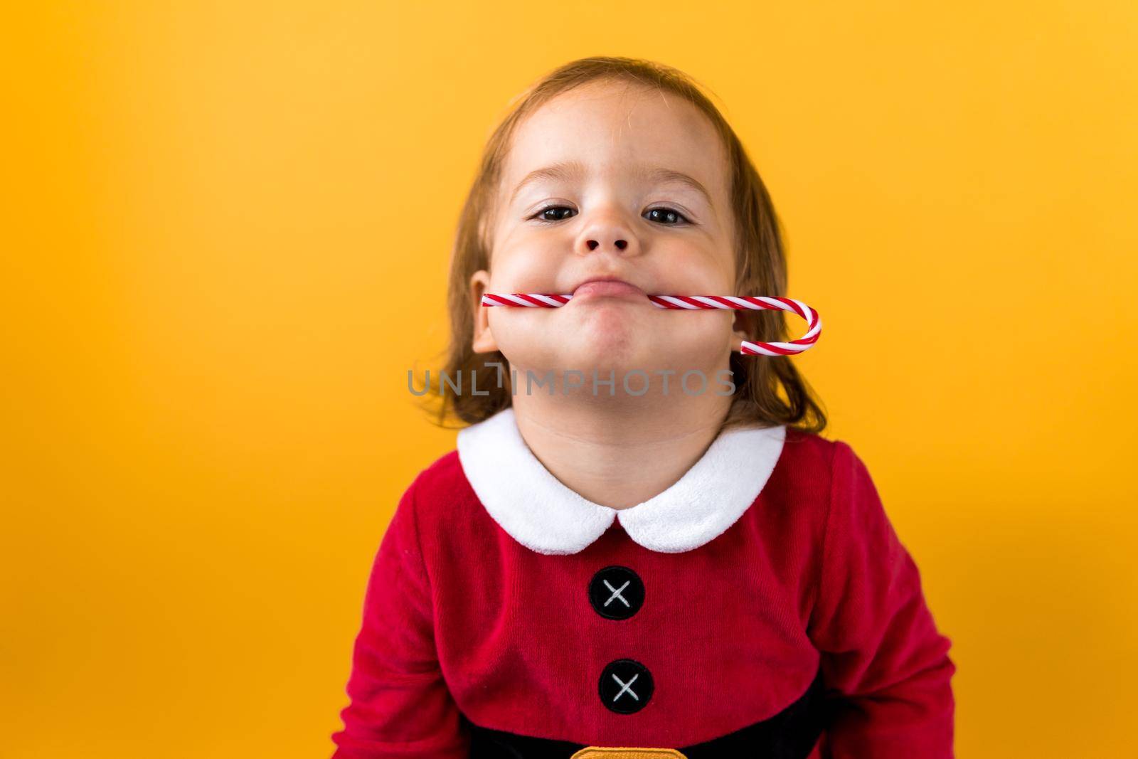 Portraite Cute Cheerful Chubby Baby Girl in Santa Suite Holding Eating Caramel Candy At Yellow Background. Child Play Christmas Scene Celebrating Birthday. Kid Have Fun Spend New Year Time Copy Space.