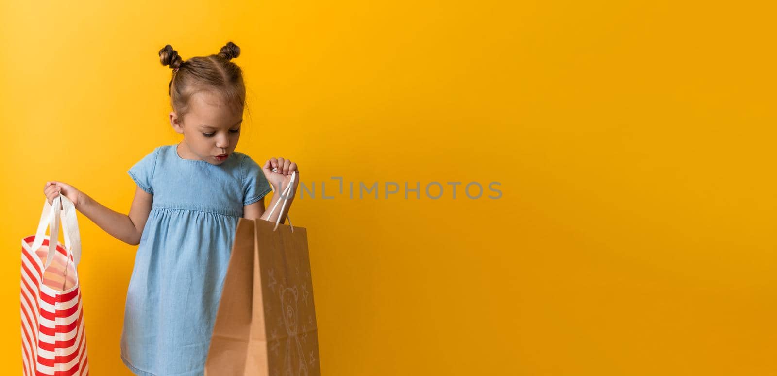 Banner Portrait Caucasian Beautiful Happy Little Preschool Girl Smiling Cheerful And Holding Cardboard Bags Isolated On Orange Yellow Background. Happiness, Consumerism, Sale People shopping Concept.