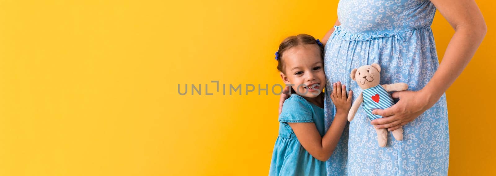 Motherhood, love, Childhood, pregnancy, hot summer - Banner pregnant unrecognizable mother woman expecting baby blue dress lillte Daughter girl sibling teddy bear hug mum belly on yellow background.